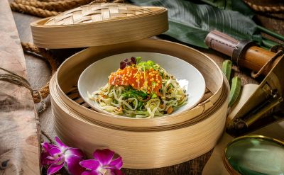 Asia Bowl im Bambuskörbchen mit gebeiztem Lachs, Wakame, Chili, Koriander und Reisperlen. Angerichtet auf einem dunklen Holztisch mit abenteuerlicher Dekoration.