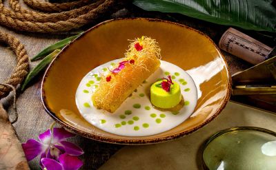 Ein Stück balinesische Ananas im Knuspermantel mit Creme aus Pandan, Honig und Pistazie. Umgeben von Orchideen und Messing-Accessoires auf dunklem Holz.