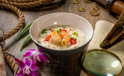 Thailändische Tom Kha Suppe mit Kokosmilch, Garnelen und Kräutern. In einer Schale angerichtet auf einem dunklen Holztisch mit abenteuerlicher Dekoration.