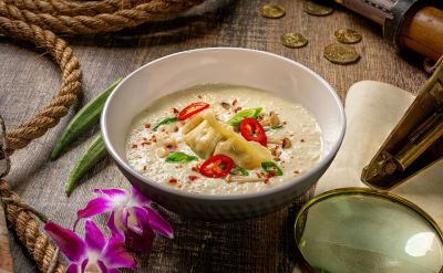 Vegane Thailändische Tom Kha Suppe mit Kokosmilch, Gemüsedumplings und Pilzen. In einer Schale angerichtet auf einem dunklen Holztisch mit abenteuerlicher Dekoration.