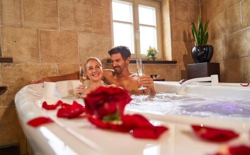 Pärchen sitzt zusammen in Badewanne, welche mit roten Rosenblättern dekoriert ist. Beide haben ein Glas Sekt in der Hand und lächeln