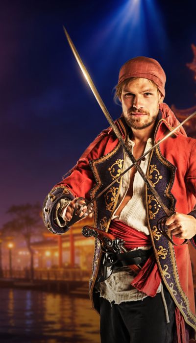 Promotionposter für die Adventure in Batavia – Immersive Dinner Experience. Ein Mann in rotem Piratenkostüm mit zwei gekreuzten Degen vor einem beleuchteten Hafen mit Segelschiff im Hintergrund.