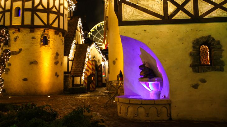 Der Froschkönig auf einem Brunnen an einer Hauswand, aus dem Brunnen scheint lila Licht