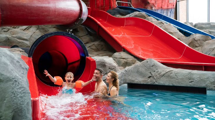 Kind kommt aus der Wasserrutsche "Översnurra" und hebt die Arme nach Oben, die Eltern warten im Schwimmbecken