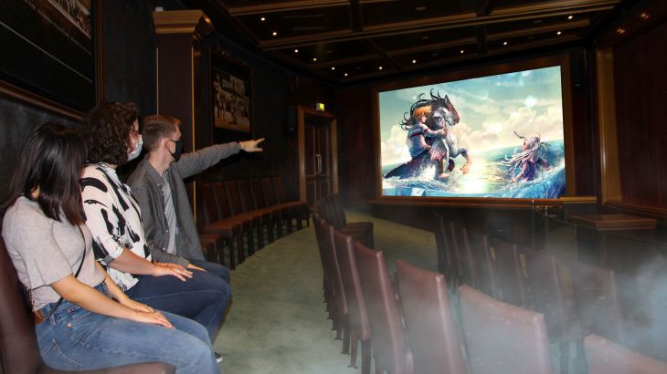 3 Leute sitzen in einem Saal, jemand zeigt auf eine Leinwand, die ein Artwork zeigt
