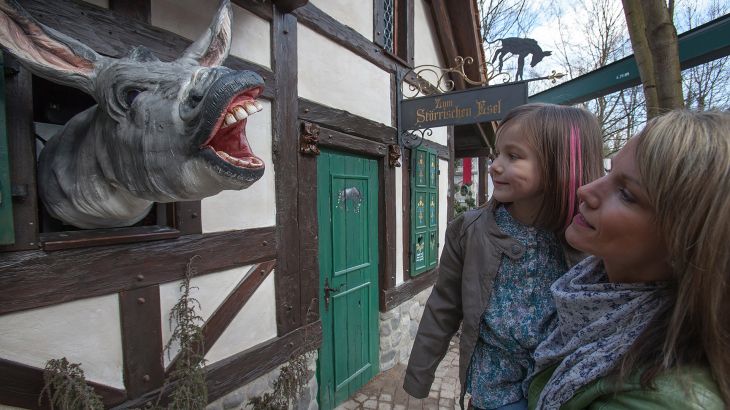 Mutter und Tochter vor einem Haus, aus welchem ein Esel aus dem Fenster schaut