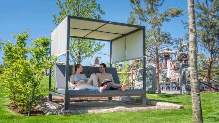 Ein Paar liegt auf dem Strand Sofa im Außenbereich von Rulantica. Im Hintergrund ist die Attraktion Svalgurok zu sehen.