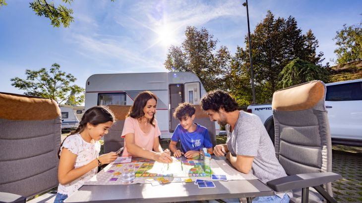 Familie sitzt vor Wohnwagen am Tisch und spielt ein Brettspiel