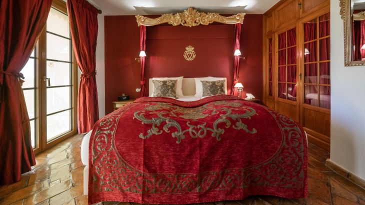 Königssuite Doppelbett mit roter Tagesdecke und goldener Verzierung links Tür zum Balkon und auf der rechten Seite ein großer Holzschrank mit roten Vorhängen