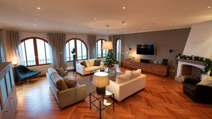 VIP-Präsidentensuite Wohnraum. Links graue Couch und rechts daneben zwei gegenüberliegende weiße Couches in der Mitte en Glastisch und dahinter drei große Panoramafenster. Auf der rechten Seite hängt ein Flachbildschirm an der Wand darunter ein Holzschrank sowie rechts daneben der Kamin