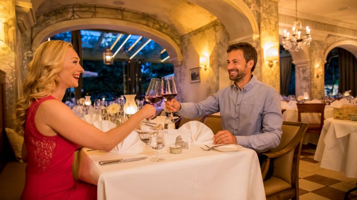 Paar sitzt im Restaurant am gedeckten Tisch, Frau links in rotem Kleid, Mann gegenüber in blauem Hemd. Beide stoßen mit einem Glas Rotwein an