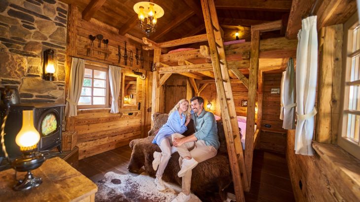 Pärchen sitzt in der Honeymoon Suite auf einer Bank vor dem Bett und lächelt. Der Innenraum der Suite ist aus Holz