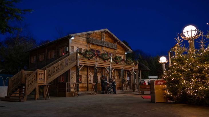 Exterior shot of the rustic ERDINGER Hütt'n at dusk. Christmas trees with fairy lights are placed in front of it.