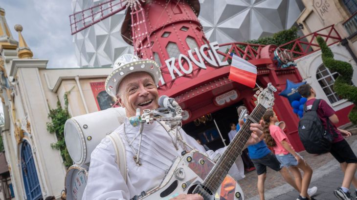 Un uomo allegro con una chitarra in mano davanti all’Eurosat - CanCan Coaster nel quartiere francese dell’Europa-Park.