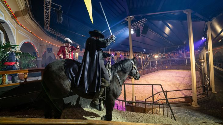 Szene aus der spanischen Arena Zorro im Europa-Park: Ein Reiter in schwarzem Umhang auf schwarzem Pferd hebt sein Schwert, ein Mann in rotem historischen Kostüm zeigt auf ihn. Überdachte Arena mit Zuschauerrängen und Fahnen.
