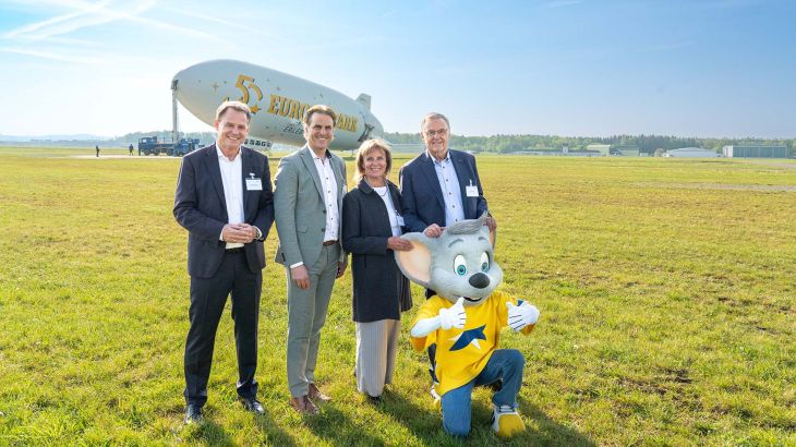 Roland, Marianne en Thomas Mack staan samen met Eckard Breuer, directeur van Zeppelin Luftschifftechnik GmbH & Co. KG, voor de Europa-Park Zeppelin. Europa-Park-mascotte Ed knielt voor hen.