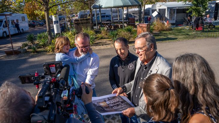 Roland Mack steht gemeinsam mit DJ BoBo und einer vierköpfigen Familie auf dem Europa-Park Caravaning und zeigt eine Illustration des "Europa-Park Kinderhaus Kleine Helden".
