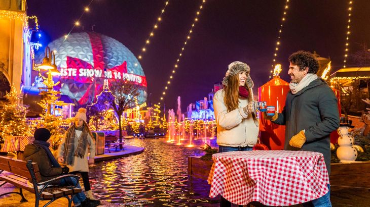 Eine Familie trinkt Heißgetränke vor der Brücke im französischen Themenbereich; im Hintergrund ist die Mapping Show auf der Eurosat Kugel zu sehen.