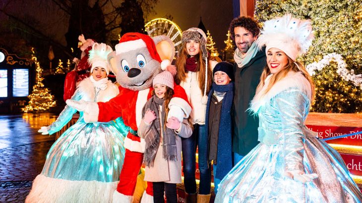 Eine Familie steht gemeinsam mit Ed Euromaus und zwei Schneeköniginnen vor einem Weihnachtsbaum.