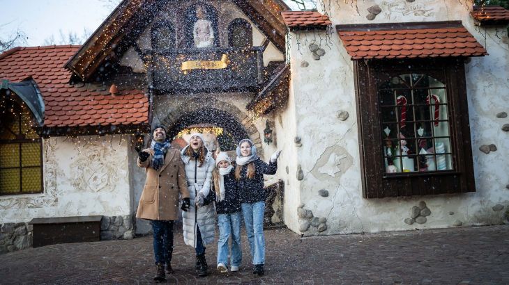 Eine vierköpfige Familie bestaunt den Schnee, welchen Frau Holle hinter ihnen aus ihren Laken schüttelt.