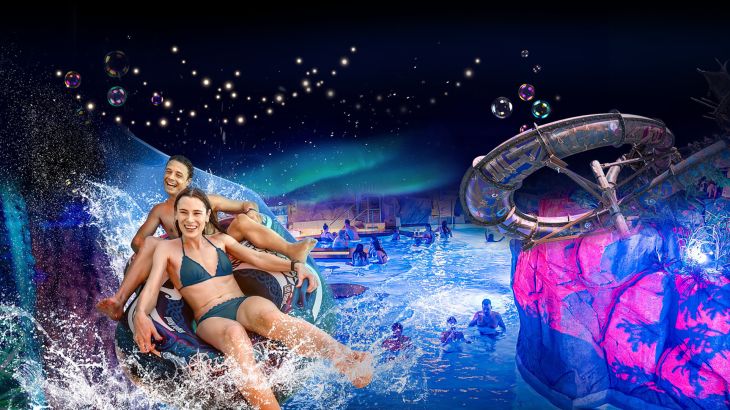 Keyvisual des Nordischen Lichterfests: Die zauberhaft beleuchtete Wasserwelt Rulantica des Europa-Park Erlebnis-Resorts. Im Vordergrund zwei Personen, die freudig eine Rutsche auf einem Reifen rutschen. 