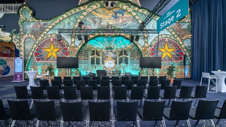 Prunkvolle Veranstaltungsbühne mit kunstvoll bemalter historischer Kulisse, dekorativen Lichtelementen und Reihen schwarzer Stühle im Vordergrund; moderne Konferenztechnik und ein Schild „Stage 2“ weisen auf einen Event- oder Kongressbereich hin.