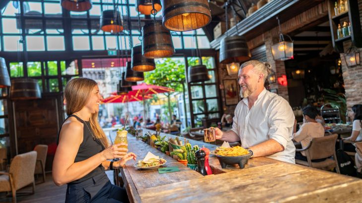 Mann und Frau sitzen sich lachend an einem Tisch gegenüber, essen gemeinsam; die Frau hält einen Cocktail.