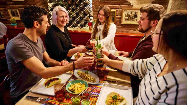 Fünf Gäste sitzen im Restaurant Don Quichotte im Erlebnishotel "El Andaluz" des Europa-Park und stoßen mit ihren Gläsern an.