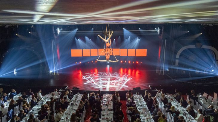 Zwei Athleten hängen an ihren artistischen Bändern in luftiger Höhe bei der Europa-Park Dinner-Show und zeigen ihre Tricks. Das Publikum sitzt an ihren Tischen und schaut gebannt zu.