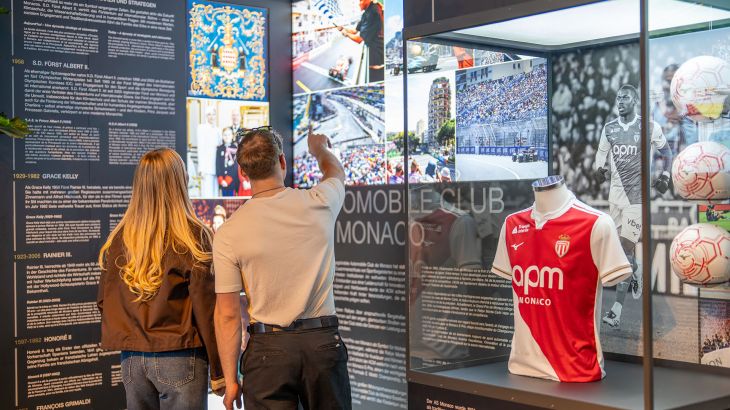 Ein Paar sieht sich die Ausstellung im Monegassischen Themenbereich im Europa-Park an. Neben dem Paar steht eine Vitrine mit einem Fußballtrikot und drei Fußbällen.