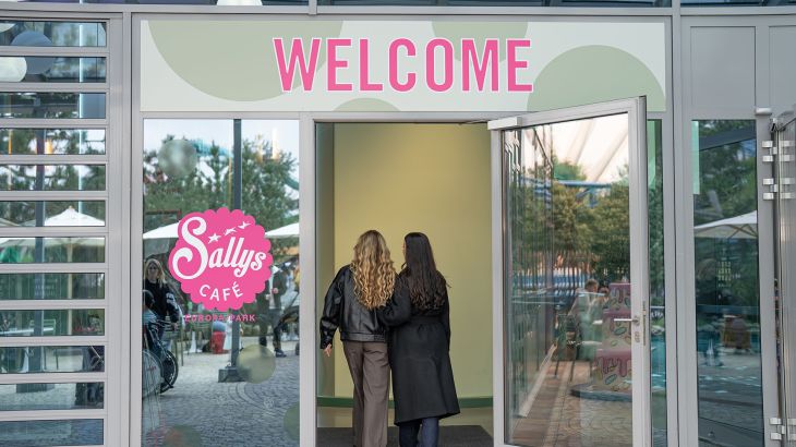 Two women walk through the entrance to Sallys Café in the Icelandic themed area of Europa-Park. The word ‘Welcome’ is written in pink letters above the door, and there is a pink logo to the left of the door.