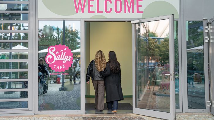 Zwei Frauen laufen durch den Eingang in Sallys Café im Isländischen Bereich des Europa-Park. Über der Türe prangt in pinker Schrift "Welcome", links neben der Tür ist ein pinkes Logo zu sehen.