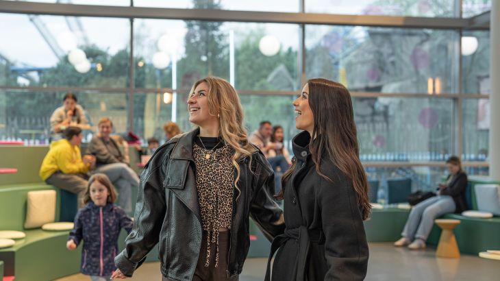 Two women are standing in the middle of Sallys Café in the Icelandic themed area of Europa-Park, laughing and looking up to the left. In the background, you can see other guests sitting in the café with their drinks.