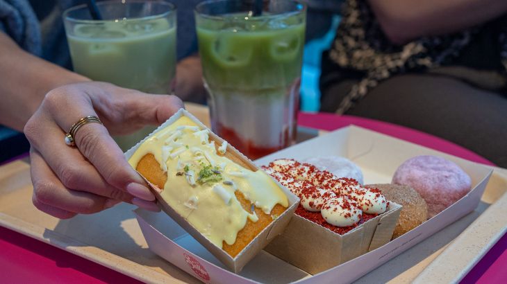 A close-up of a tray with three mini doughnuts, two small cakes and two iced strawberry matcha lattes at Sallys Café in the Icelandic themed area of Europa-Park. A hand is holding one of the small cakes.