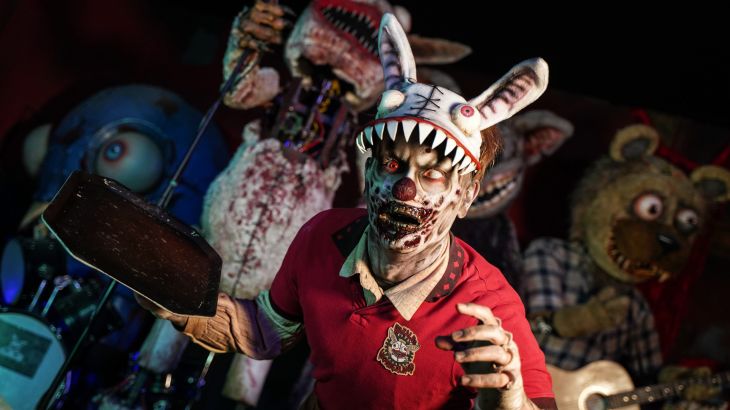 A character from Traumatica dressed as a zombie, wearing a creepy rabbit hat and holding a tray. In the background, you can see other horror figures.