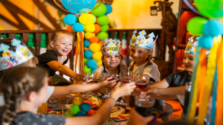 Sechs Kinder stoßen beim Kindergeburtstag im Europa-Park mit Saftgläsern an. Vier davon tragen einen JUNIOR CLUB Partyhut.