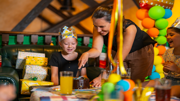 Mädchen mit JUNIOR CLUB Partyhut am gedeckten Tisch beim Kindergeburtstag im Europa-Park. Frau schneidet Schokoladenkuchen an.