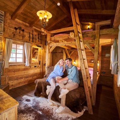 Pärchen sitzt in der Honeymoon Suite auf einer Bank vor dem Bett und lächelt. Der Innenraum der Suite ist aus Holz