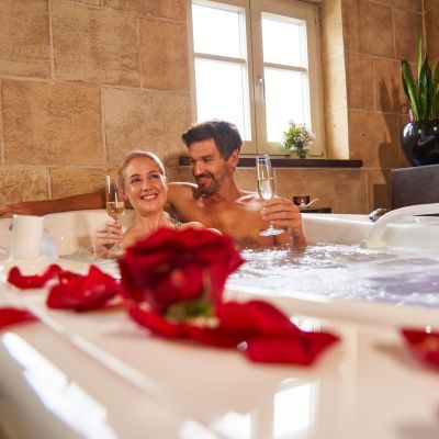 Pärchen sitzt zusammen in Badewanne, welche mit roten Rosenblättern dekoriert ist. Beide haben ein Glas Sekt in der Hand und lächeln
