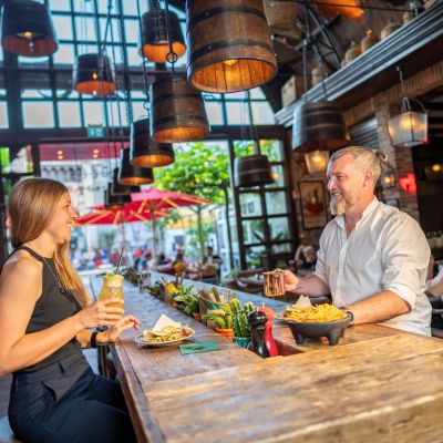 Mann und Frau sitzen sich lachend an einem Tisch gegenüber, essen gemeinsam; die Frau hält einen Cocktail.