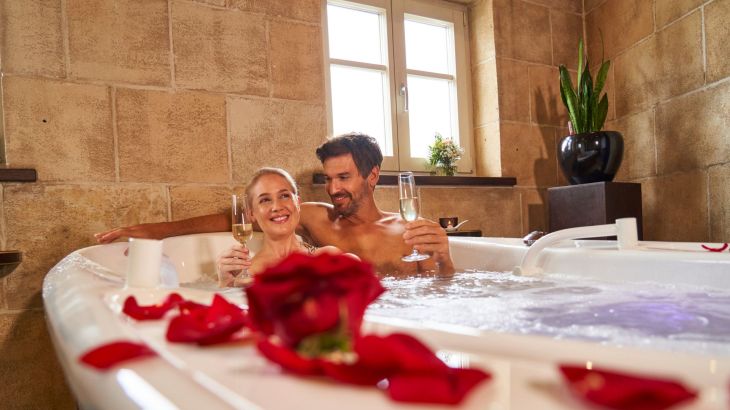 Pärchen sitzt zusammen in Badewanne, welche mit roten Rosenblättern dekoriert ist. Beide haben ein Glas Sekt in der Hand und lächeln