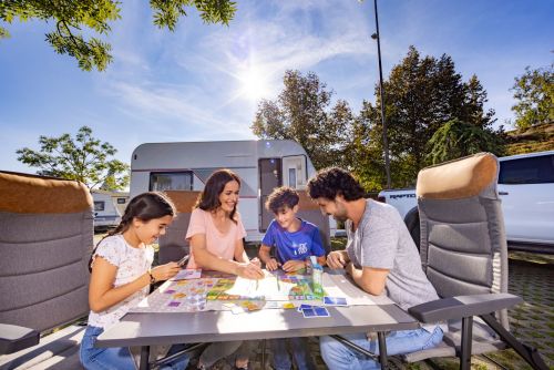 Familie sitzt vor Wohnwagen am Tisch und spielt ein Brettspiel