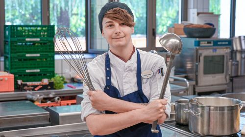 eine Köchin mit gekreuzten Armen, in der einen Hand eine Schöpfkelle, in der anderen Hand ein Schneebesen.