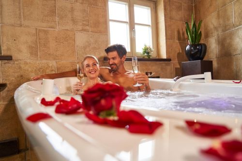 Pärchen sitzt zusammen in Badewanne, welche mit roten Rosenblättern dekoriert ist. Beide haben ein Glas Sekt in der Hand und lächeln