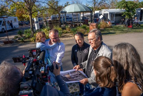 Roland Mack steht gemeinsam mit DJ BoBo und einer vierköpfigen Familie auf dem Europa-Park Caravaning und zeigt eine Illustration des "Europa-Park Kinderhaus Kleine Helden".
