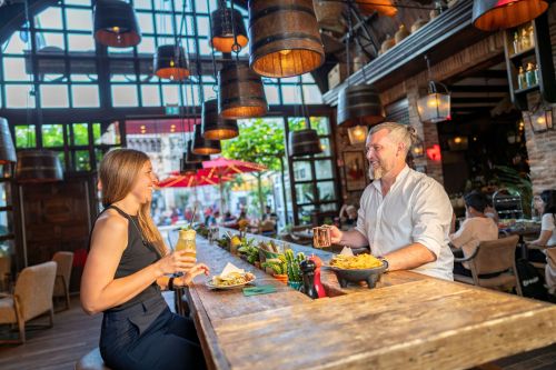 Mann und Frau sitzen sich lachend an einem Tisch gegenüber, essen gemeinsam; die Frau hält einen Cocktail.