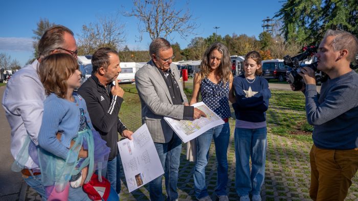 Roland Mack demonstriert die Pläne des "Europa-Park Kinderhaus Kleine Helden" vor laufender Kamera, neben ihm DJ BoBo und eine vierköpfige Familie.
