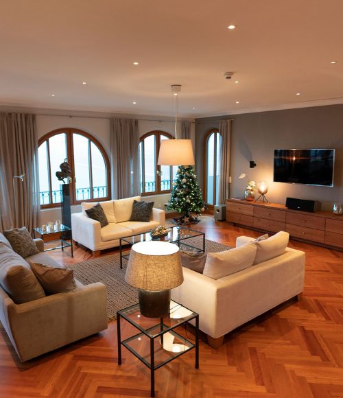 VIP-Präsidentensuite Wohnraum. Links graue Couch und rechts daneben zwei gegenüberliegende weiße Couches in der Mitte en Glastisch und dahinter drei große Panoramafenster. Auf der rechten Seite hängt ein Flachbildschirm an der Wand darunter ein Holzschrank sowie rechts daneben der Kamin