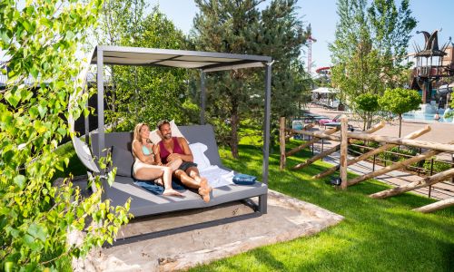 Ein Paar liegt auf einem Strand Sofa im Außenbereich von Rulantica. Im Hintergrund ist die Attraktion Svalgurok zu sehen.