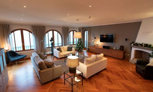 VIP-Präsidentensuite Wohnraum. Links graue Couch und rechts daneben zwei gegenüberliegende weiße Couches in der Mitte en Glastisch und dahinter drei große Panoramafenster. Auf der rechten Seite hängt ein Flachbildschirm an der Wand darunter ein Holzschrank sowie rechts daneben der Kamin